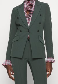 Zelené dvouřadé sako s černými knoflíky, které má dvě přední kapsy a lehce kostkované volánkové manžety vyčnívající z pod rukávů.