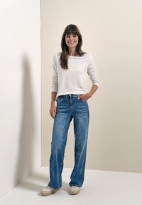 Femme aux longs cheveux bruns, portant une chemise blanche à manches longues, un jean bleu et des baskets blanches, debout avec une main dans la poche et souriante.