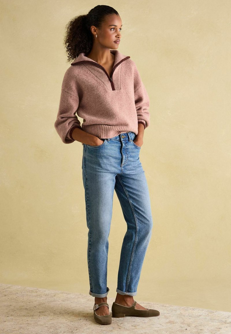 Hellrosa gestrickter Pullover mit Reißverschlusskragen, kombiniert mit hochgeschnittenen blauen Jeans und braunen flachen Schuhen. Weiche Texturen und lockere Passform.