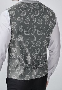 Grau gemusterte Weste mit floralen Motiven und weißen Umrandungen. Verfügt über ein Rückband-Detail und wird über einem weißen Hemd getragen. Schlichte schwarze Hose.