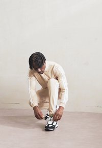 Chándal beige con rayas blancas, persona agachada atándose zapatillas en blanco y negro sobre un suelo gris claro contra un fondo neutro.