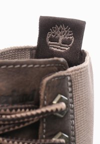 Botín de cuero marrón con una parte superior de tela beige, que presenta un diseño cosido, ojales hexagonales y una etiqueta de tela marrón con un logo.