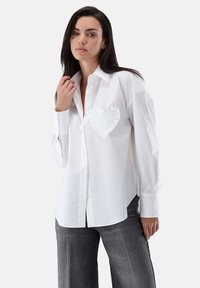White button-up shirt made of cotton with a ruffled heart patch on the left chest, featuring long sleeves and a tailored fit.