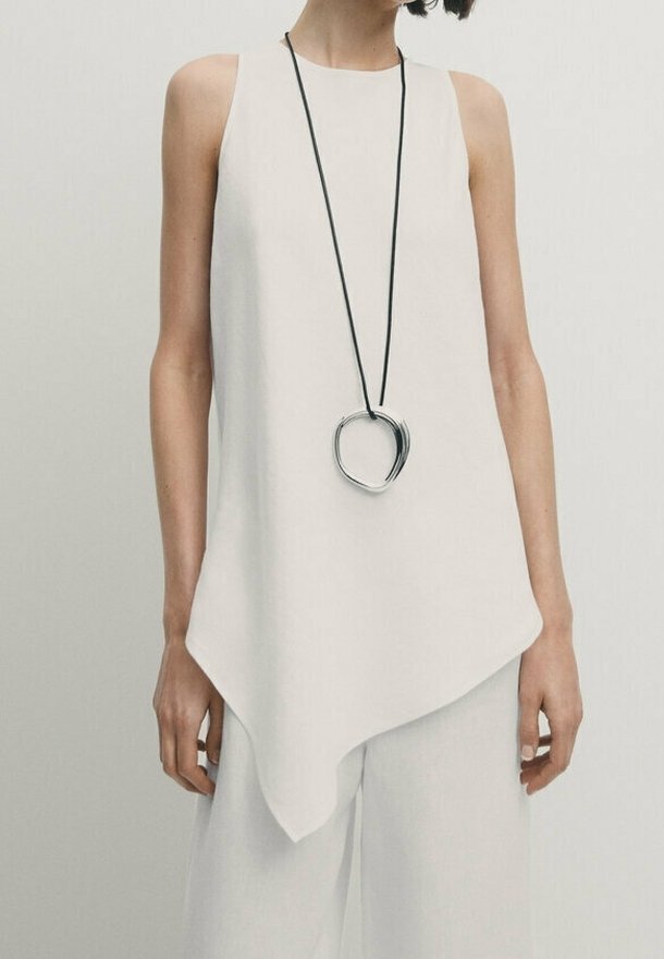 Femme portant un haut blanc asymétrique sans manches avec un pantalon blanc à jambes larges et un long collier noir avec un grand pendentif circulaire en argent.