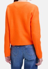 Kvinna som bär en livlig orange långärmad fluffig tröja och blå denimjeans, sedd bakifrån mot en vanlig bakgrund.