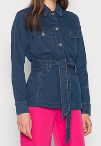 Jeansjacka med midjebälte, två bröstfickor och metallknappar. Mörkblå färg med kontrasterande sömmar. Långa ärmar med knäppta manschetter.