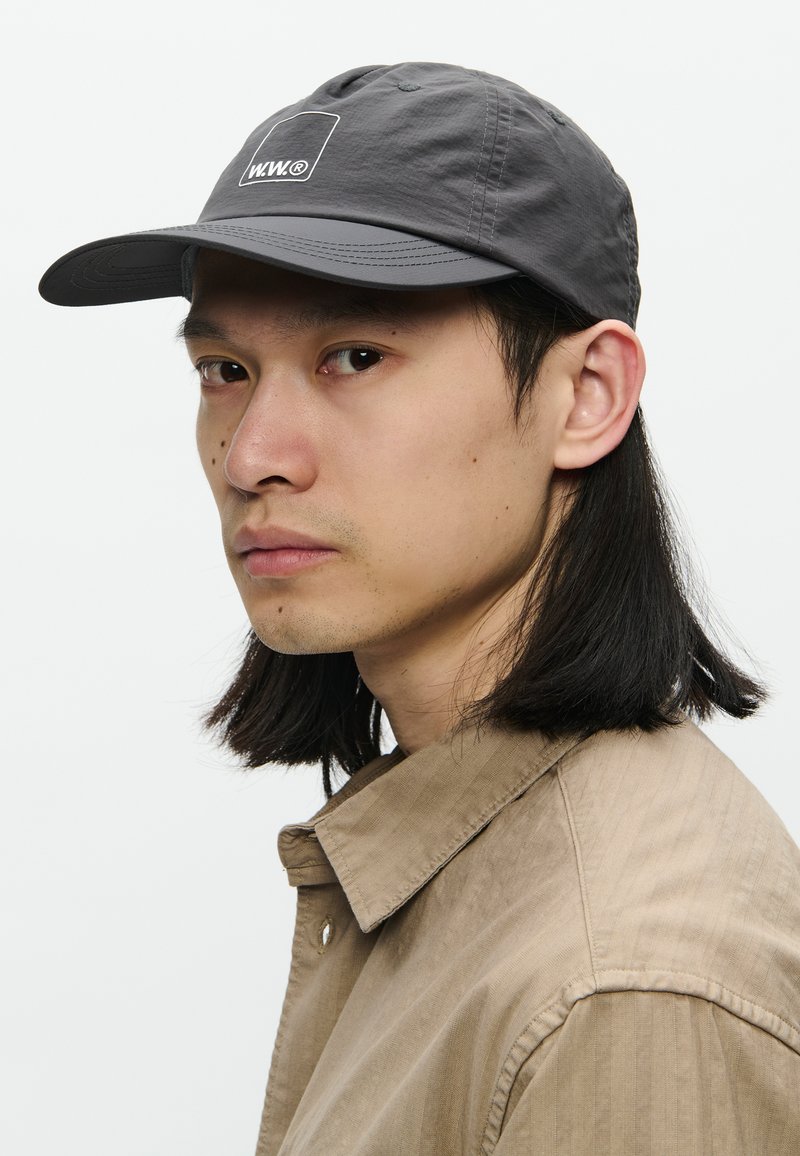 Giovane uomo con capelli neri lunghi fino alle spalle, che indossa un cappello grigio con il logo "W.W®" e una camicia beige con colletto, che guarda leggermente di lato.