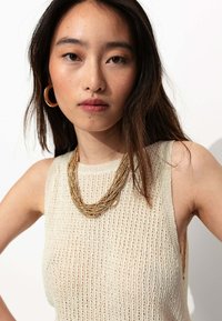 Pull sans manches en tricot blanc cassé avec un motif texturé, associé à un collier multibrins en ton or et de grandes boucles d'oreilles en or.