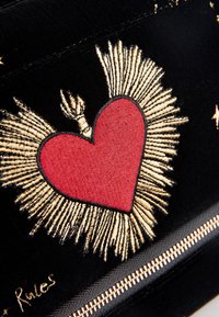 Schwarze Stofftasche mit einem roten Herzmotiv, verziert mit goldenen Fadenakzenten und Strahlen. Goldener Reißverschluss mit schwarzem Rand entlang der unteren Kante.