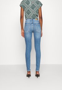 Jeans ajustados de mezclilla azul claro con cintura alta, dos bolsillos traseros y una sutil textura desgastada. Combinados con una blusa verde con estampado.