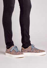 Patterned sneakers featuring a textured brown and blue floral design, white rubber soles, and light blue laces, paired with dark jeans.