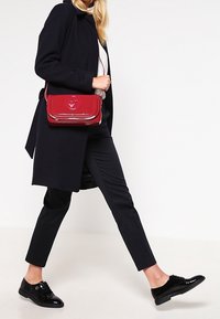 Persona che cammina indossando un cappotto nero e pantaloni neri, scarpe lucide nere, portando una borsa a tracolla rossa con un logo sul risvolto anteriore.