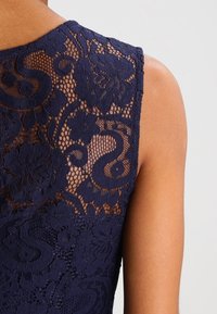 Navy blue lace top featuring intricate floral patterns and a textured design, with a sleeveless cut and sheer back detail.