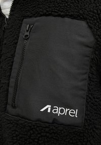 Giacca in pile nera con tasca con zip, caratterizzata da un tessuto nero liscio e logo "aprel" bianco, che evidenzia i contrasti di texture e funzionalità.