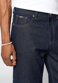 Tummansiniset farkut, joissa on keltainen ommel, viisi taskua ja tukevat vyötärö. Kuvassa näkyy käsi, jossa on helmihihna.