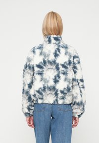 Chaqueta de piel sintética en un patrón de tie-dye azul y blanco, diseño corto, cuello alto y puños elásticos, combinada con pantalones de mezclilla azules.