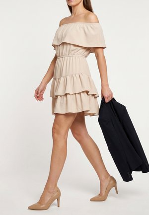 Kobieta w beżowej sukience z odkrytymi ramionami i falbankami oraz beżowych szpilkach idzie, trzymając czarną marynarkę w prawej ręce.