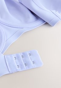 Bretelle de soutien-gorge violet clair avec trois crochets métalliques et boucles en tissu sur un fond beige lisse, montrant les détails de la fermeture.