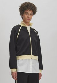 Zwarte zip-up jas met gele accenten en hoge kraag. Heeft lange mouwen en een getailleerde taille, gelaagd over een wit shirt.