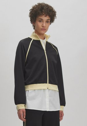Schwarze Zip-Jacke mit gelben Akzenten und hohem Kragen. Verfügt über lange Ärmel und eine taillierte Passform, die über einem weißen Hemd getragen wird.