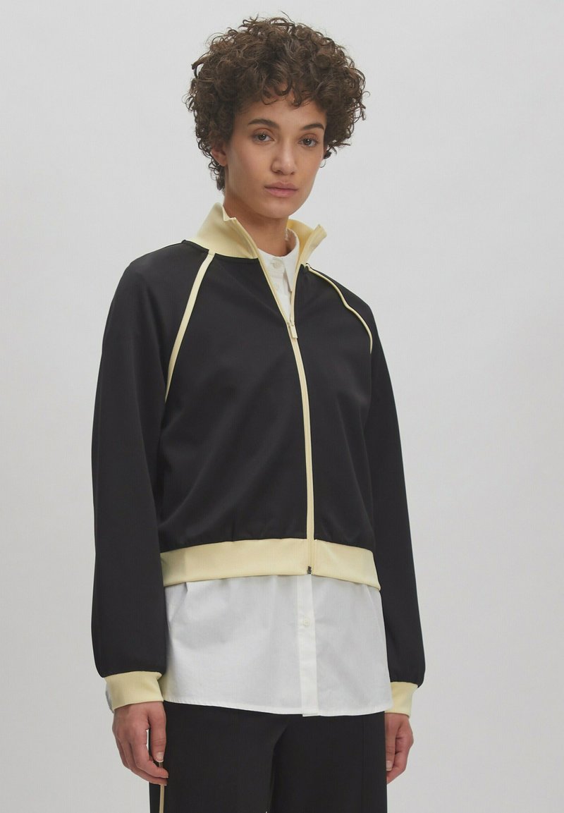 Zwarte zip-up jas met gele accenten en hoge kraag. Heeft lange mouwen en een getailleerde taille, gelaagd over een wit shirt.
