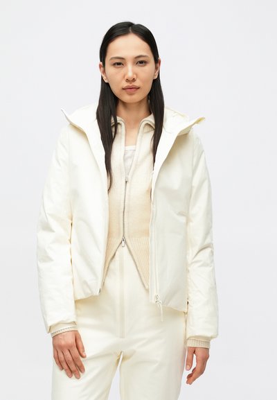 Veste matelassée blanche et isolante avec capuche, dotée d'une fermeture éclair intégrale et de poignets élastiques. Portée sur un pull beige texturé.