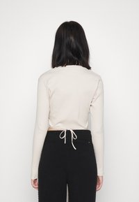 Kvinna med axellångt mörkt hår som bär en beige långärmad topp knuten bak och svarta högmidjade byxor, stående mot en vit vägg.