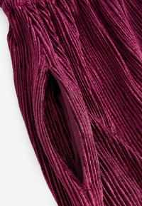 Burgundy corduroypohkeet, joissa on pystyridgid ja kudottu rakenne, sekä piilotettu tasku sivussa lisäämään toiminnallisuutta.