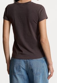 Person som bär en enkel mörkbrun t-shirt med korta ärmar och ljusblå denimjeans, sedd bakifrån mot en neutral bakgrund.