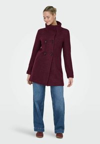 Burgundijas vilnas mētelis ar divkāršo pogu dizainu, lielām melnām pogām, augstu apkakli un slaidu piegriezumu, kombinēts ar vaļīgām zilām džinsām.