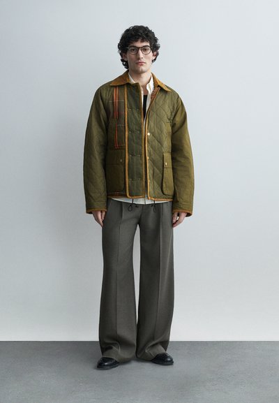 Νέος άνδρας με γυαλιά, φορώντας φούρνιστο μπουφάν ελιάς με καφέ περιθώριο, λευκό πουκάμισο, φαρδιά γκρι παντελόνια και μαύρα παπούτσια, στέκεται μπροστά σε γκρι τοίχο.