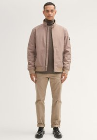Lichtroze bomberjack met een hoge kraag, beige gebreide trui eronder, gecombineerd met lichtbruine broek en zwarte schoenen. Vlotte textuur, minimalistisch ontwerp.
