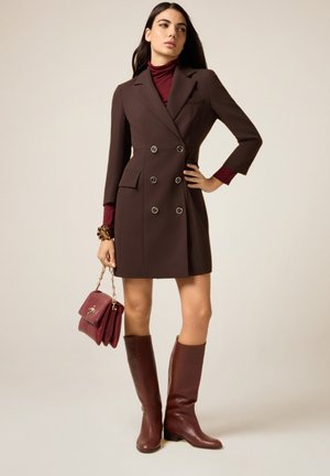 Donna che indossa un vestito blazer doppiopetto marrone, dolcevita color marrone scuro, stivali abbinati al ginocchio, che tiene una borsa marrone scuro con catena dorata.