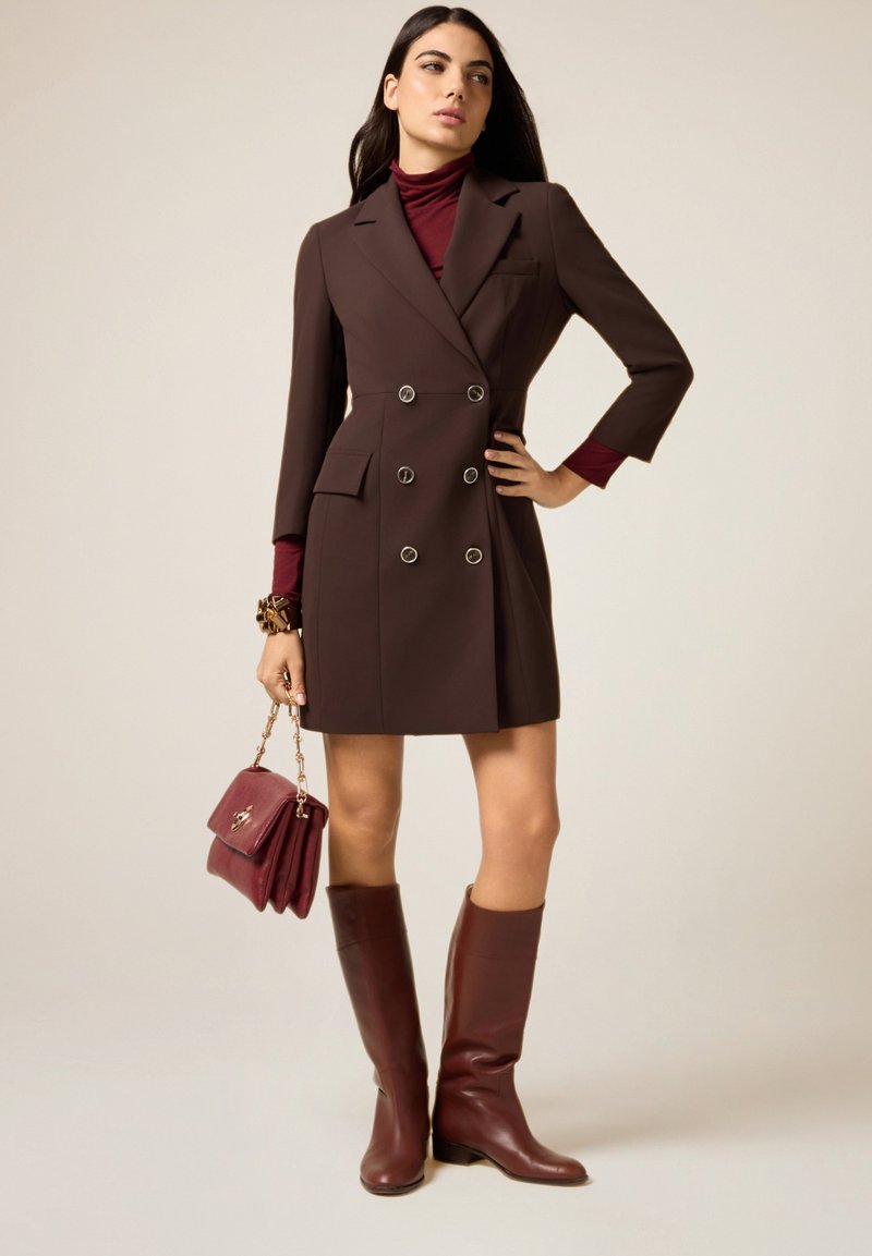 Donna che indossa un vestito blazer doppiopetto marrone, dolcevita color marrone scuro, stivali abbinati al ginocchio, che tiene una borsa marrone scuro con catena dorata.