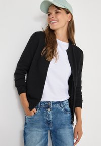 Černý pletený cardigan s žebrovanými manžetami, nošený přes bílé tričko a s lehkými modrými džíny. Modelka má také na sobě mátovou kšiltovku.