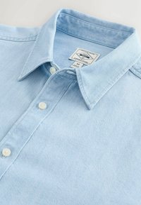 Chemise en jean bleu clair boutonnée avec col pointu et étiquette de marque visible à l'intérieur du col, taille moyenne.