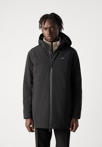Veste imperméable noire à manches longues avec capuche, dotée d'une fermeture éclair à l'avant et d'un logo sur la poitrine. Texture lisse et design minimaliste.