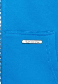Blaue Kapuzenjacke mit strukturierter Stoffoberfläche, ausgestattet mit einem silbernen Logoetikett mit der Aufschrift "bazix republic" auf der Tasche.