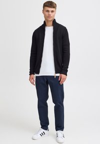 Svart stickad cardigan med ribbad krage, bärs över en vit skjorta. Matchas med marinblå byxor och vita sneakers med svarta ränder.