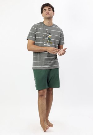 Hombre de pie descalzo, vestido con una camiseta de rayas grises con un gráfico de McDuck y pantalones cortos verde oscuro, sobre un fondo blanco.