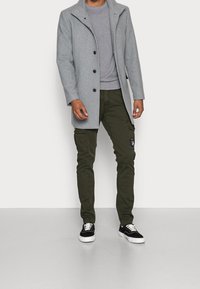 Manteau en mélange de laine grise avec un col montant et cinq boutons, associé à un pull gris chiné et un pantalon cargo vert foncé. Chaussures noires.