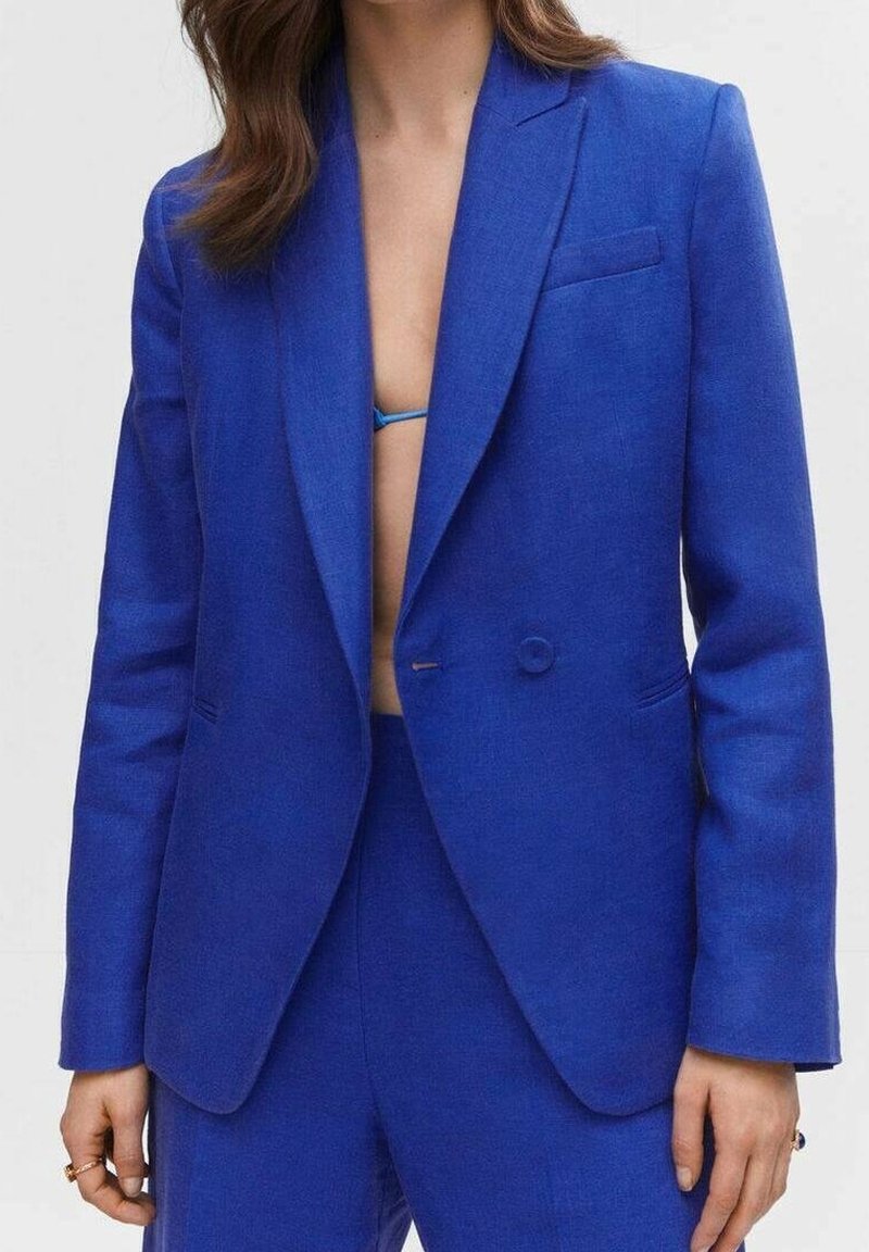 Femme portant un blazer bleu vif sur mesure et un pantalon assorti, avec un haut de bikini bleu visible en dessous, debout devant un fond uni.