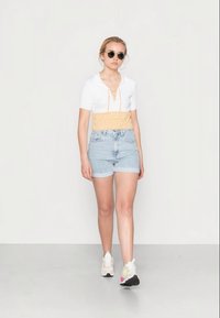 Top branco canelado de mangas curtas com um cós franzido amarelo, combinado com calções de ganga claros e ténis brancos; modelo a usar óculos de sol redondos.