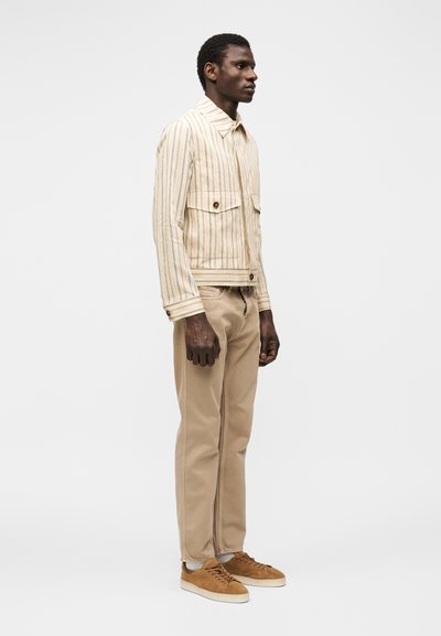 Homme debout de profil portant une veste beige rayée, un pantalon beige clair, des chaussettes blanches et des baskets en daim marron sur fond blanc.