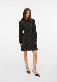 Robe noire à motifs floraux avec des manches longues, un col boutonné, une taille cintrée et une jupe évasée. Associée à des escarpins bruns à bout pointu.