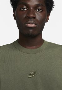 Tamsiai žalia medvilninė marškinėliai su apvali apykakle ir siuvinėtą Nike logotipą derančia žalia spalva ant krūtinės, su tekstūruotu raišteliu.