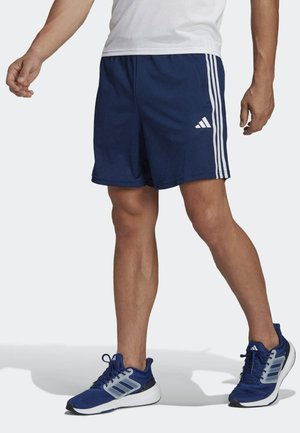 Homme portant un short Adidas bleu marine avec des bandes blanches et des baskets Adidas bleues assorties, se tenant sur un fond neutre.