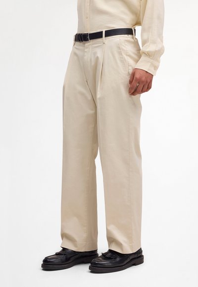 Homme portant un pantalon beige plissé, ceinture noire, chemise crème à manches longues, et mocassins en cuir noir, debout devant un fond uni.