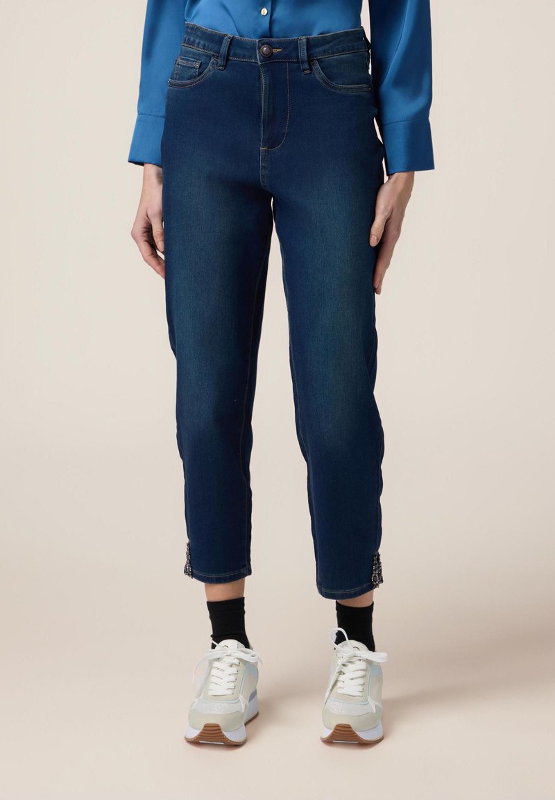 Jeans de mezclilla azul oscuro con un diseño ajustado, que cuentan con un cierre de botón, bolsillos frontales y detalles decorativos de encaje en el tobillo.
