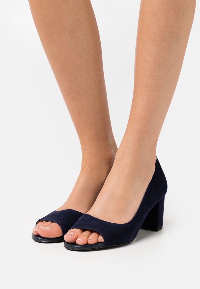 Jambes portant des chaussures à talons larges en daim bleu marine à bout ouvert sur un fond blanc uni.